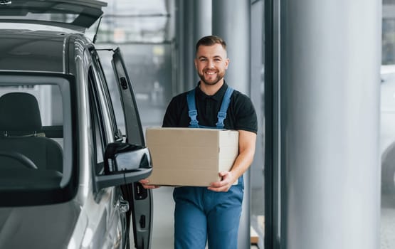 In blue colored clothes. Delivery man in uniform is indoors with car and with order.