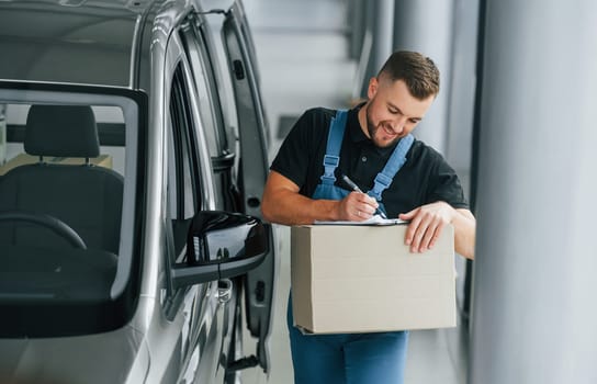 In blue colored clothes. Delivery man in uniform is indoors with car and with order.