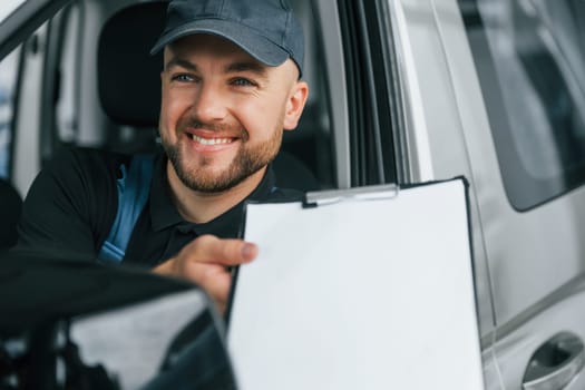 Cheerful worker. Delivery man in uniform is indoors with car and with order.