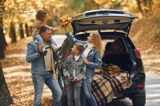 Near car with open trunk. Happy family is in the park at autumn time together.