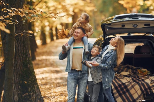 Near car with open trunk. Happy family is in the park at autumn time together.