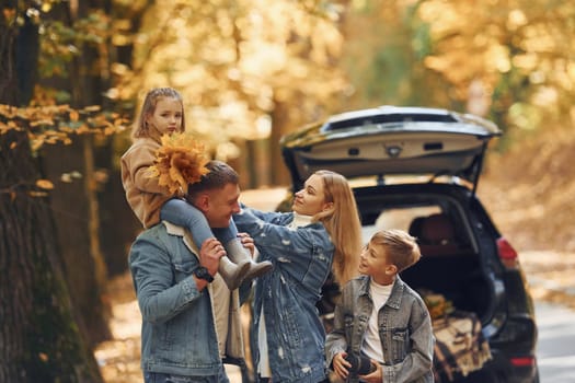 Near car with open trunk. Happy family is in the park at autumn time together.