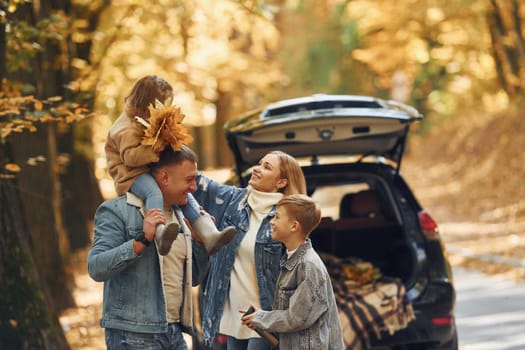 Near car with open trunk. Happy family is in the park at autumn time together.