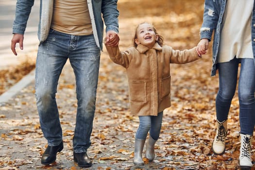 Holding daughter by hands. Happy family is in the park at autumn time together.