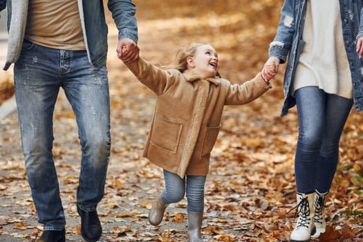 Holding daughter by hands. Happy family is in the park at autumn time together.