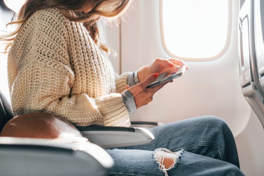 Sitting with phone in hands. Young female passanger in casual clothes is in the plane.