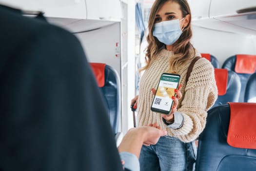 Showing COVID-19 vaccine sertificate to stewardess. Young female passanger in casual clothes is in the plane.