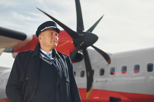 Pilot in formal black uniform is standing outdoors near plane.