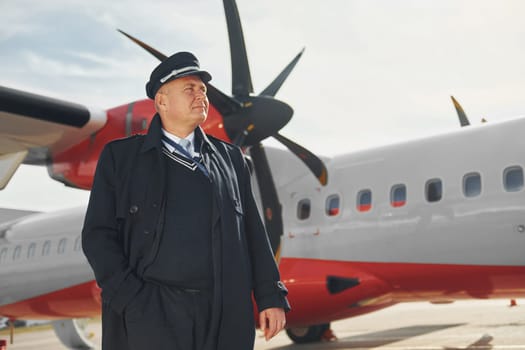Pilot in formal black uniform is standing outdoors near plane.