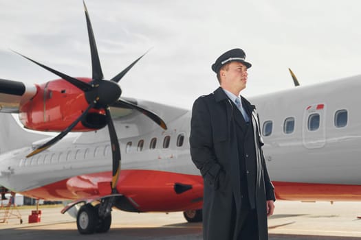 Looking far away. Pilot in formal black uniform is standing outdoors near plane.