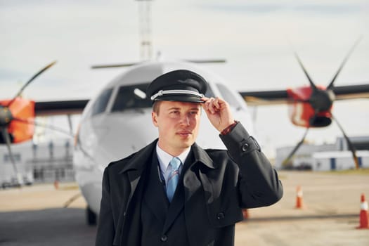 Pilot in formal black uniform is standing outdoors near plane.