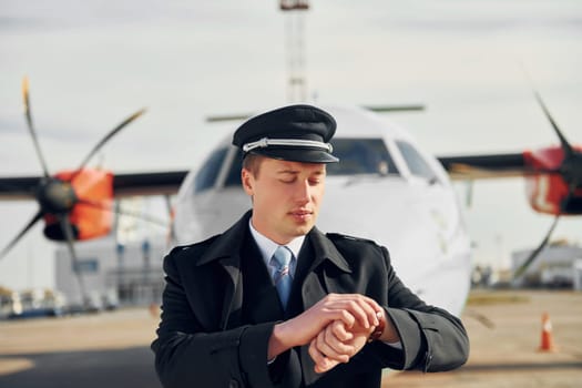 Checking the time. Pilot in formal black uniform is standing outdoors near plane.