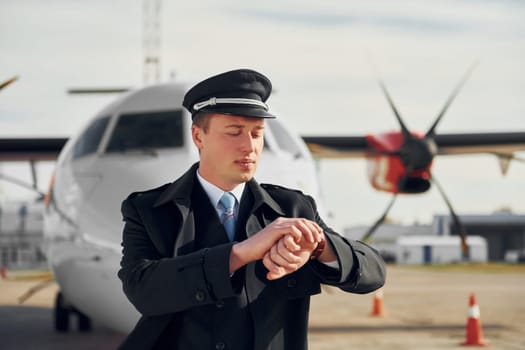 Checking the time. Pilot in formal black uniform is standing outdoors near plane.