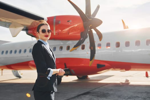 Young stewardess that is in formal black clothes is standing outdoors near plane.