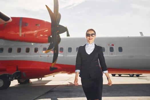 Looking far away. Young stewardess that is in formal black clothes is standing outdoors near plane.
