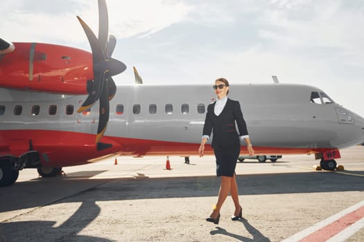 Looking far away. Young stewardess that is in formal black clothes is standing outdoors near plane.