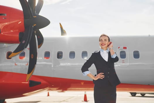 Looking far away. Young stewardess that is in formal black clothes is standing outdoors near plane.