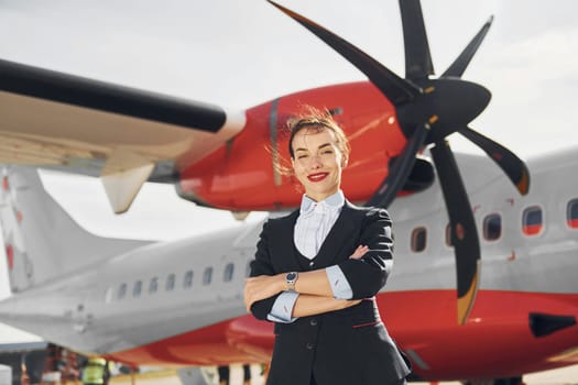 At daytime. Young stewardess that is in formal black clothes is standing outdoors near plane.