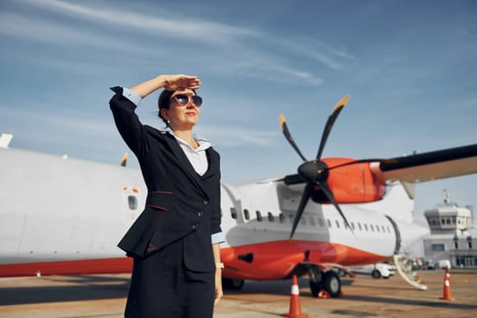 At daytime. Young stewardess that is in formal black clothes is standing outdoors near plane.