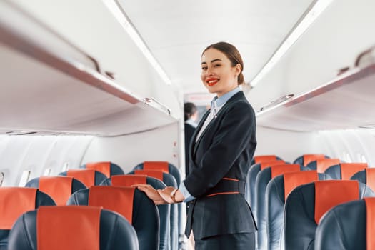 Young stewardess that is in formal black clothes is standing indoors in the plane.