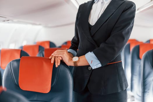 Young stewardess that is in formal black clothes is standing indoors in the plane.