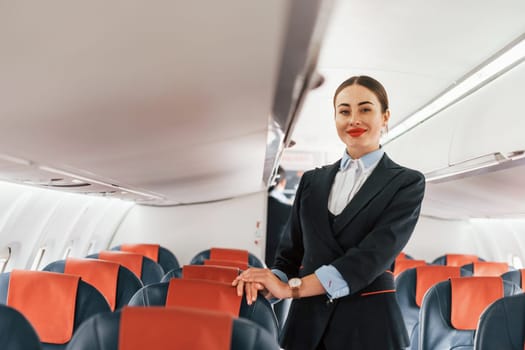 Young stewardess that is in formal black clothes is standing indoors in the plane.