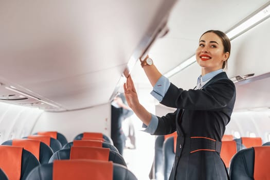 Young stewardess that is in formal black clothes is standing indoors in the plane.