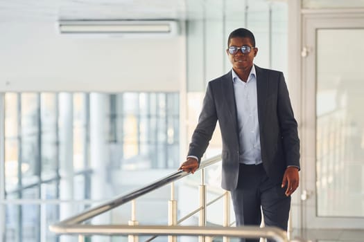 Leaning on the railings. Young african american businessman in black suit is indoors.