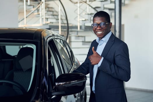Standing near car. Young african american businessman in black suit is the autosalon.