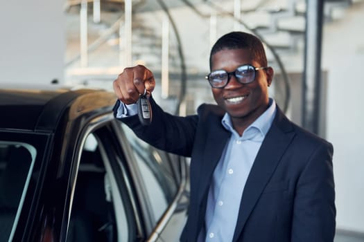 Standing near car. Young african american businessman in black suit is the autosalon.