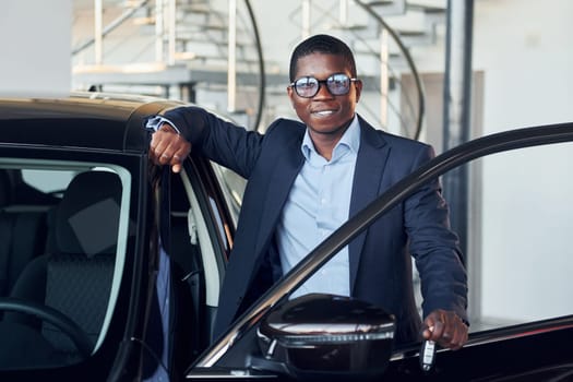 Holding keys in hands. Young african american businessman in black suit is the autosalon.