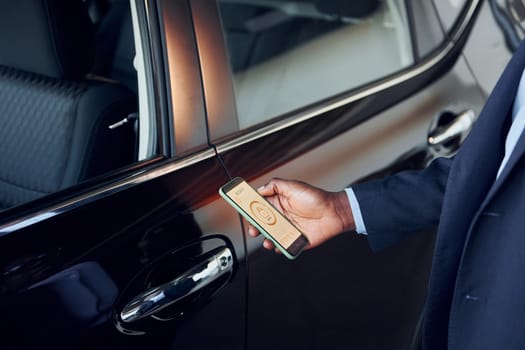 Using smartphone to open door. Young african american businessman in black suit is the autosalon.
