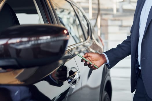 Using smartphone to open door. Young african american businessman in black suit is the autosalon.