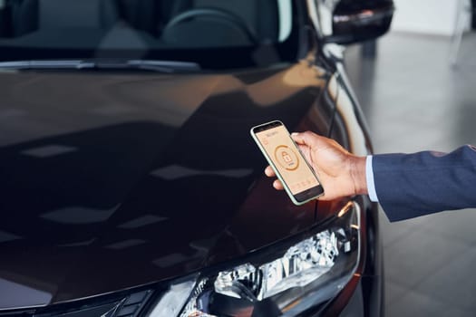 Using smartphone to open door. Young african american businessman in black suit is the autosalon.