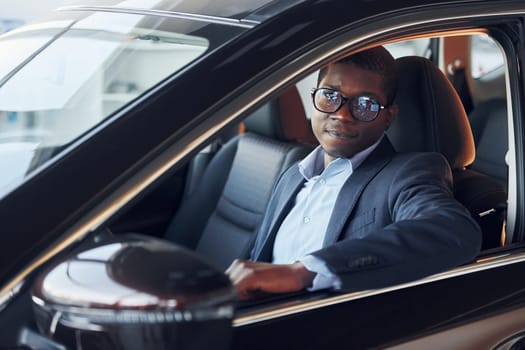 Positive mood. Young african american businessman in black suit is in the automobile.