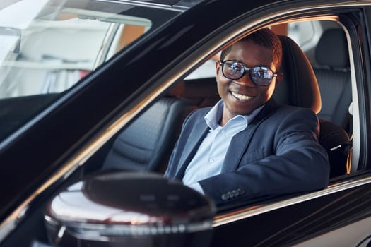 Positive mood. Young african american businessman in black suit is in the automobile.