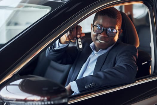 Positive mood. Young african american businessman in black suit is in the automobile.