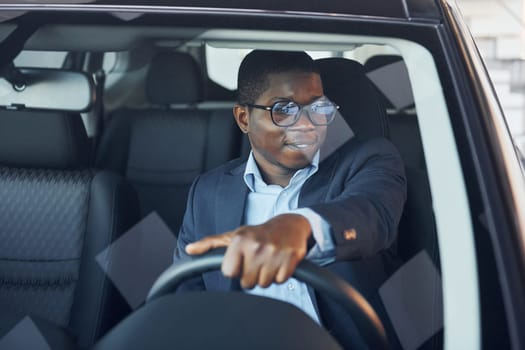 Front view. Young african american businessman in black suit is in the automobile.
