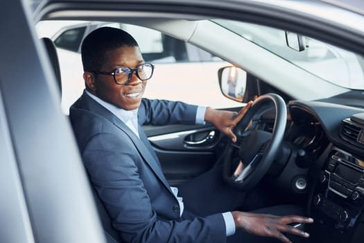 Side view. Young african american businessman in black suit is in the automobile.