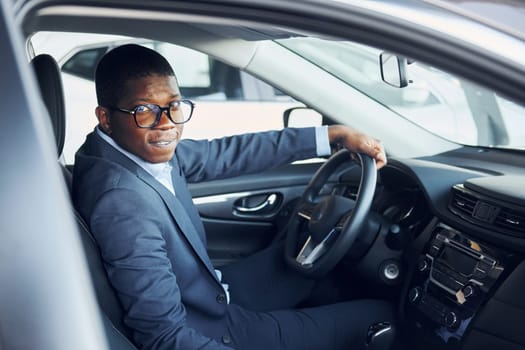 Side view. Young african american businessman in black suit is in the automobile.