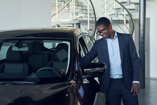 Looking at car. Young african american businessman in black suit is the autosalon.