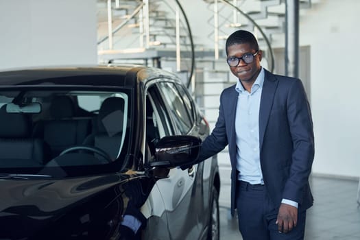Looking at car. Young african american businessman in black suit is the autosalon.