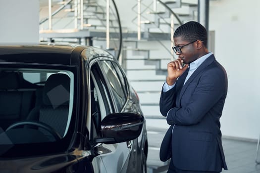 Looking at car. Young african american businessman in black suit is the autosalon.