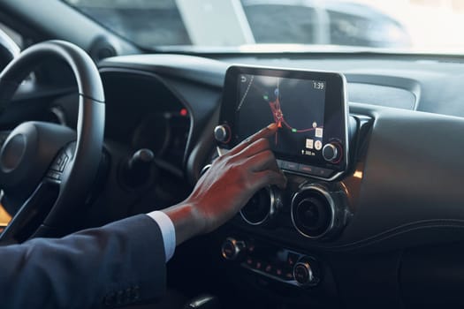 Using front control panel. Young african american businessman in black suit is in the automobile.