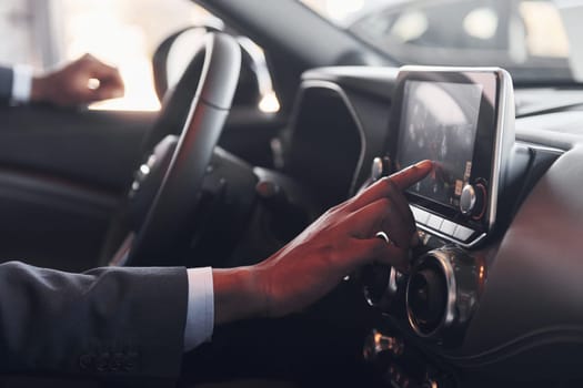 Using front control panel. Young african american businessman in black suit is in the automobile.