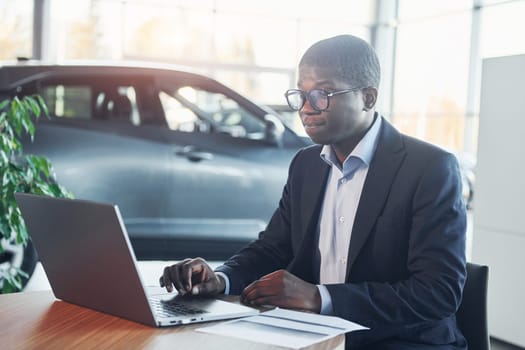 Office worker is sitting in autosalon by table with laptop.