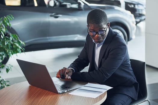 Office worker is sitting in autosalon by table with laptop.