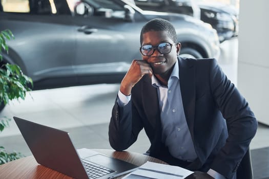 In glasses. Office worker is sitting in autosalon by table with laptop.