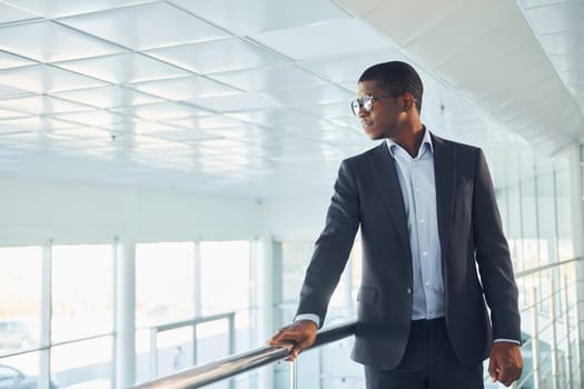 Leaning on the railings. Young african american businessman in black suit is indoors.