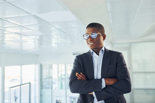 With arms crossed. Young african american businessman in black suit is indoors.